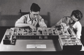 Black-and-white photo of two museum preparators sitting at a table while they work on an exhibit.
