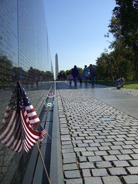 panoramic view of memorial wall -- a small American flag rests against the memorial wall in the foreground; the Washington Monument appears in the background