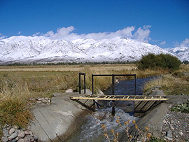 wooden bridge spanning irrigation canal in foreground; snow-covered mountains and blue sky in background.