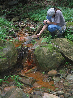 female in baseball cap leaning over rust-colored stream.
