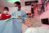 Two medical personnel wearing surgical masks attend a patient in a hospital bed.