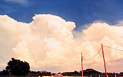 Large, light-colored clouds billowing  in the background, with blue sky above.  A farm is in the foreground.