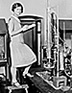 Photo: woman, standing on a footstool, with laboratory instruments.