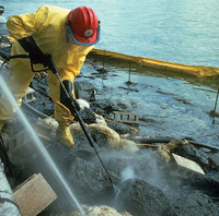 Worker using power washer to spray oil off rocks on a beach.