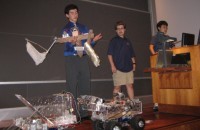 three students presenting their robot in the final competition; one student is holding the robot and other robotic vehicles are on the floor