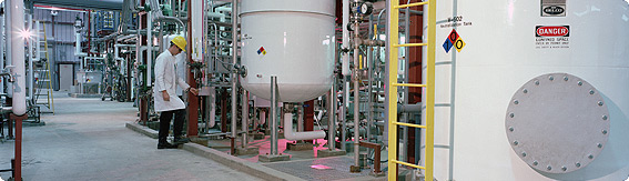 Photo of a long hallway with two large neutralization tanks.  The large, cylindrical tanks are surrounded by a network of tubes, and a ladder stands against the closer of the two tanks.  A man in a lab coat and a safety helmet is checking the tubes at the farther of the tanks.