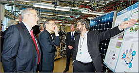 In photo, two men look on as a scientist explains his photovoltaics research using a large poster with scientific diagrams.