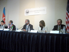 Panel of five people in suits at a table with microphones. The American and Russian flags are behind them. Each are drinking bottled water.