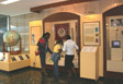 State Department Exhibit hall -- Three people standing in front of a glass-enclosed alcove containing artifacts. To the left is a large world globe encased in a glass showcase. State Department photo.