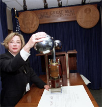 Sharon Bisdee standing inside glass enclosure of the Great Seal, with hand on Great Seal press after impressing a document
