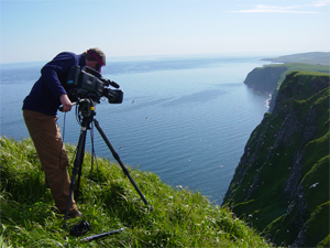 a man operating a video camera on a cliff over looking the sea.