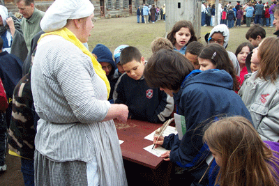 a lady in a blue and white striped jacket and yellow scarf stands with a group of children over a table.