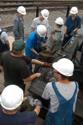 high school students wearing white hardhats discuss railroad equipment with one of steamtown's mechanics.