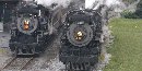 Two steam locomotives pulling passenger trains pass by the loading platform at Steamtown.