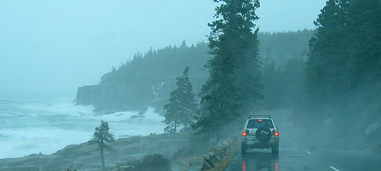 Waves pound the coastline while a car sits on the road in rainy weather.