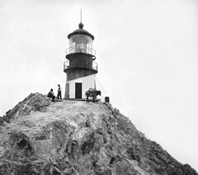 A historic photo of the lighthouse on the Farallon Islands with two men and a mule next to it.