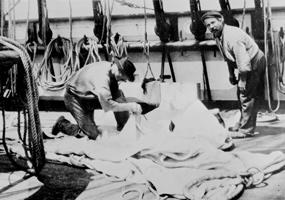 A sailmaker and his assistant repairing a sail laying on the deck of a ship.