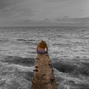 photo of a person sitting at the end of a narrow wooden dock, surrounded by grey waves under a grey sky