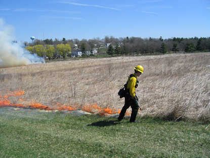 A firefighter in yellow gear ignites the dry grasses of the prairie with a drip torch.