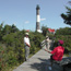 One person sits on a bench while others walk on a boardwalk trail with the black-and-white banded Fire Island Lighthouse in the background.