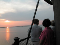 Standing along the black iron railing, several people gaze at pink and orange reflections in the water as the sun sets over the Great South Bay.