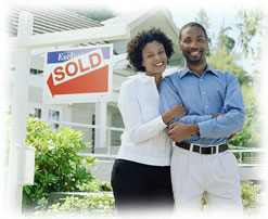 Photograph of a couple standing smiling next to a sold sign in front of a house.