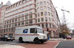 Image 1: A USPS Mail truck drives down a busy city street.