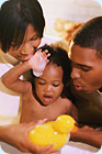 A mother and father giving their baby a bath; mom is holding a rubber duckie.