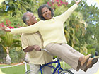 An older woman, laughing, riding on the handle bars of a bike; an older man riding the bike and smiling at her.