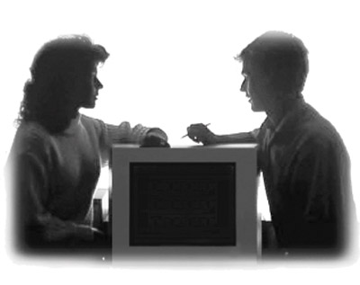 Side view photo of a young male and female sitting across from one another with a computer monitor between them.