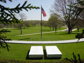 Two plain marble slabs and a U.S. flag on a green lawn framed by spruce trees.