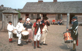 a group of people stand with drums and fifes in lines, on a dirt area, playing music