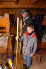 A child in a winter jacket stands in a soldier's barracks passing wooden muskets from a continental soldier to other children.