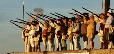 Soldiers stand on the walls of the fort at sunset, muskets leveled at an unseen target. Grey smoke lingers in clumps surrounding them.