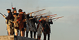 men stand in formation on the corner of a fort wall as they level their muskets, smoke billows around them
