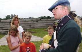 A tall soldier wears a green woolen jacket, he is speaking to children in multicolored tee-shirts on a corner of the fort.