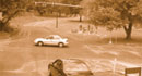 This photograph shows an intersection in a rural setting. A traffic light hangs over an intersection of two roads. It is red. One car is in the intersection and another car in the cross-traffic road sits waiting at the light.