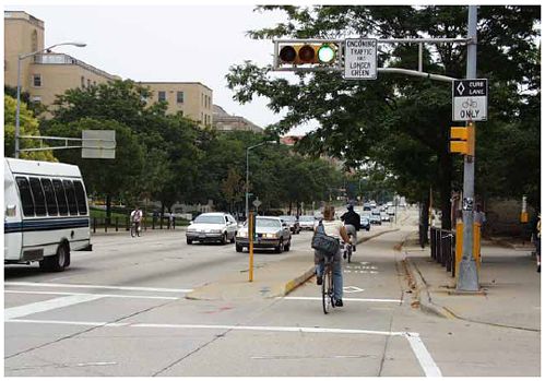 This photograph shows a contraflow lane that runs counter to the motor vehicle traffic lanes. The contraflow bike lane is separated from the vehicle lanes by a concrete divider, and a special signal is used for the bike lane. A sign next to the bike signal says -ONCOMING TRAFFIC HAS LONGER GREEN.