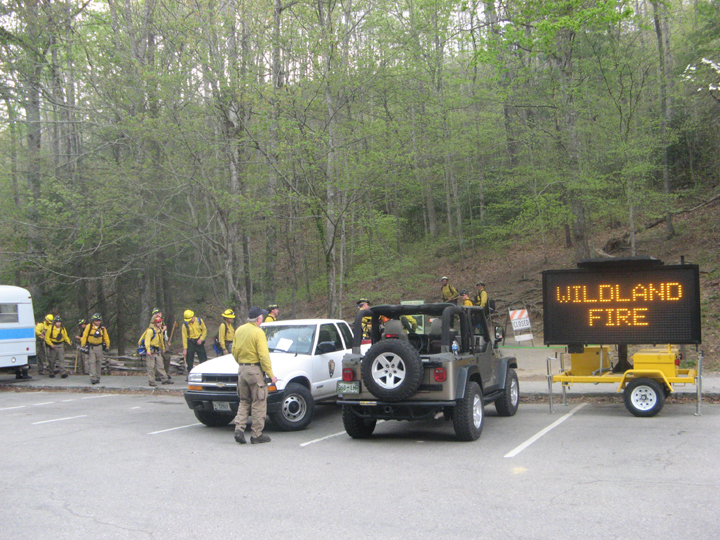 photo: A group of firefighters walks from a gray schoolbus to a trailhead.  An electronic sign in the parking lot says, 