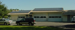 Photo of long, 1-story building with 2 garage doors. Black solar panels cover roof.