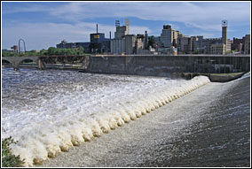 The urban skyline of Minneapolis looms over the tamed St. Anthony waterfall.  Water spills over a large concrete apron.