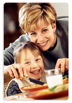 smiling adult woman helping smiling girl eat meal at table with a variety of food on a plate and milk in a glass.