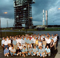 This image shows about 50 people standing and kneeling together as they pose outside in front of the tall Delta II rocket that perches toward the sky about 1/2 mile in the distance.  The sky is grey with total cloud coverage, and the rocket base blends in with the blue-grey clouds.  The top of the rocket and four visible rocket boosters on the bottom shine in white.  In between the people and the rocket stands a large, black steal scaffolding structure that looks somewhat like the Eiffel Tower, without the pointy peak.