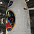 In this image, in the center, is the large white, protective fairing bearing both the red, white and blue NASA 'meatball' logo and the very colorful mission logo.  The fairing sits atop the large Atlas V rocket that will launch it out of Earth's atmosphere, toward Mars.  The inside of the vertical integration facility is very industrial looking with large steel beams, tubing and service stairways.  There is a tube attached to the fairing that provides air conditioning to the spacecraft.