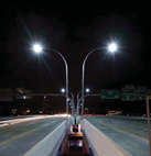 View at night looking down a bridge made up of two sections.  Between the sections is a base holding two bright street lamps, shaped liked antennas, the left one curved to the left and the right one curved to the right.