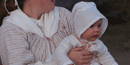 A small baby is held in its mother's arms. It is wearing a white cap, and a white fabric sheet as a covering. Its mother clutches it tightly
