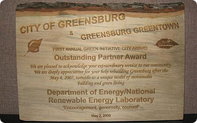 Photo of a rectangular wooden plaque with writing on the facing surface. The top of the plaque bears a layer of bark from the tree from which it was made. The plaque reads, in part, City of Greensburg & Greensburg GreenTown First Annual Green Initiative City Award Outstanding Partner Award … Department of Energy/National Renewable Energy Laboratory "Encouragement, generosity, counsel"