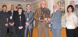 Photograph:  Logan Lee (far left), Gene DeGayner (2nd left), Dave Schmid (3rd left; behind Anne), Anne Zimmermann (4th left; front), Tim Dabney (5th left),Corbin Newman and Don DeLorenzo (center; holding award), Liz Agpaoa (2nd from right) and Tom Peterson (far right, behind Liz).