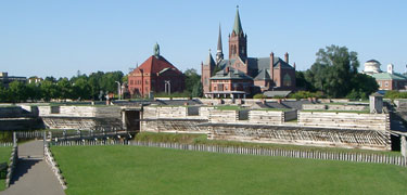 Blue skies above, a green lawn below, in between a wooden fort rises, with old churches and red brick city buildings even taller behind.