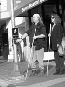 A man and a woman are standing at the edge of the street with stores visible behind them. Each holds a long white cane. The male has his right arm extended, with his hand on a pushbutton on a pole to his right side. 
